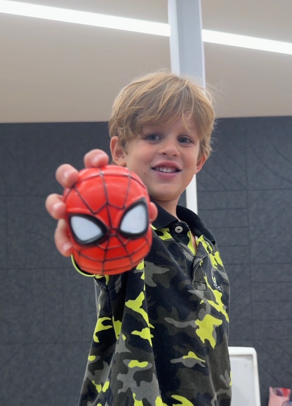 A smiling child holding a red Spider-Man Mojis squishy toward the camera.